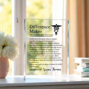 May include: A clear rectangular glass plaque featuring black printed text that defines a Difference Maker as a physical therapist. The design includes a PT caduceus symbol and the name Susan Thomas at the bottom, presented on a flat surface.
