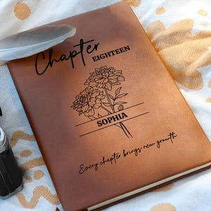 Könnte beinhalten: Ein braunes Lederjournal mit den Worten "Chapter Eighteen" und "Sophia" auf dem Einband eingraviert. Ein Blumendesign und der Satz "Every chapter brings new growth" sind ebenfalls abgebildet. Eine Feder liegt auf der oberen linken Ecke.