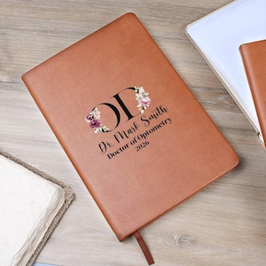 May include: A brown leather-bound journal with the text "Dr. Mark Smith, Doctor of Optometry 2026" and the initials "OD" decorated with floral accents. A brown ribbon bookmark is visible.