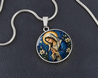 Puede incluir: Un collar con colgante circular de tono plateado que presenta una ilustración detallada de una figura con un halo dorado, vistiendo una túnica y un velo azul, con las manos juntas en oración entre lirios blancos sobre un fondo azul estrellado.