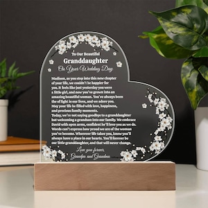 May include: A heart-shaped acrylic plaque with a wooden base, featuring white floral accents and a heartfelt message for a granddaughter on her wedding day. The text includes a personal message from her grandparents.