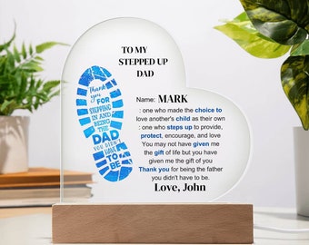 May include: A blue and white heart-shaped acrylic plaque with a blue footprint design and the text "Thank you for stepping in and being the Dad you didn't have to be." The plaque also includes the text "To my Stepped Up Dad" and "Name: Mark" followed by a definition of what it means to be a stepped up dad. The plaque is on a wooden base and has a light behind it.