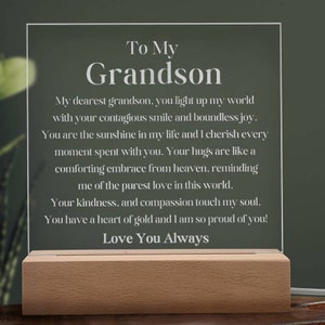 May include: A white acrylic plaque with a wooden base, displaying a heartfelt message to a grandson. The message reads: "To My Grandson, My dearest grandson, you light up my world with your contagious smile and boundless joy. You are the sunshine in my life and I cherish every moment spent with you. Your hugs are like a comforting embrace from heaven, reminding me of the purest love in this world. Your kindness, and compassion touch my soul. You have a heart of gold and I am so proud of you! Love You Always."