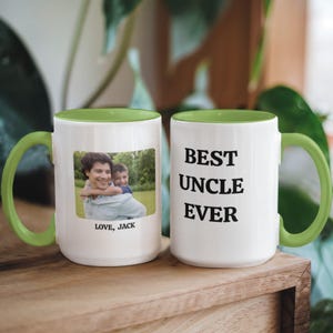 May include: Two white ceramic mugs with green handles and green interiors. One mug has a photo of a man and child with the text "LOVE, JACK". The other mug has the text "BEST UNCLE EVER" in black.