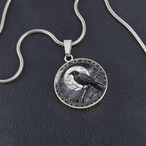May include: A silver-toned circular pendant necklace featuring a black and white illustration of a raven perched on a branch against a full moon. The pendant is suspended from a matching silver-toned snake chain with a polished metallic finish.
