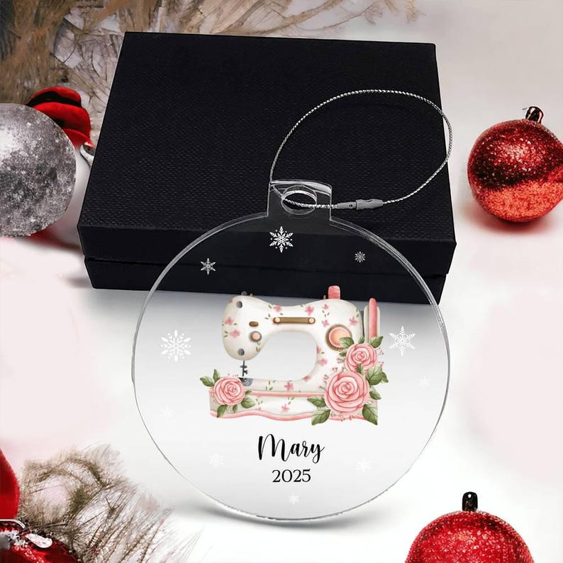 May include: Clear, round Christmas ornament featuring a sewing machine design with pink roses and snowflakes. The ornament has the name "Mary" and the year "2025" printed on it. A silver wire loop is attached for hanging. The ornament is displayed with red and silver Christmas decorations.