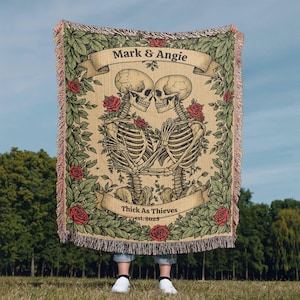 May include: A woven tapestry throw blanket with a floral design featuring two skeletons holding each other. The text "Mark & Angie Thick As Thieves est. 2023" is printed on the blanket.