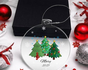 May include: A clear, round Christmas ornament with three decorated green Christmas trees. The ornament has a silver hanging wire and the name "Mary" and the year "2025" printed on it. The ornament is surrounded by red and silver holiday decorations.