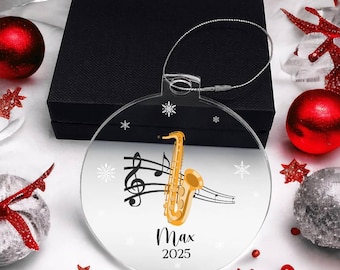 May include: A clear, circular acrylic ornament featuring a gold saxophone illustration, black musical staff lines, and white snowflake accents. The text "Max" and "2025" is printed in black. A silver hanging cord is attached to the top of the ornament.