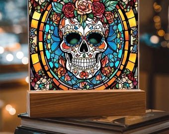 May include: A decorative square light featuring a sugar skull design with floral accents. The skull is white with intricate details, surrounded by a stained-glass effect with vibrant colors. It sits on a wooden base, perfect for home decor.
