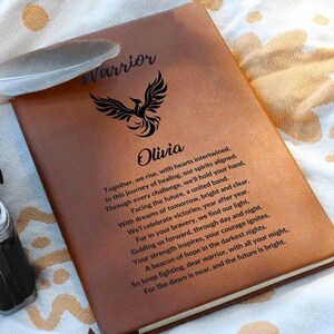 May include: A brown leather journal with the word "Warrior" and a phoenix design engraved on the front. The name "Olivia" is also engraved on the front. The journal has an inspirational quote engraved on the front.