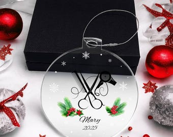 May include: Clear, round Christmas ornament with a salon theme. The ornament features scissors, a comb, and a hairdryer. The name "Mary" and the year "2025" are printed on the ornament. The ornament is surrounded by snowflakes, red berries, and green sprigs.