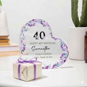 May include: A heart-shaped clear acrylic plaque decorated with purple floral patterns, featuring the text 40, HAPPY 40TH BIRTHDAY, Samantha, Forty & Fabulous, Love, Sarah, & Alex, accompanied by a small square gift box tied with a purple ribbon.