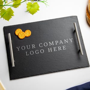 May include: Black slate serving tray with two silver handles. The tray has the text "YOUR COMPANY LOGO HERE" etched into the surface. There are three crackers on the tray.