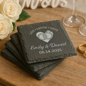 May include: Set of four black slate coasters with a white engraved design. The design features two interlocking rings inside a heart with the text "Live Laugh Love" above and "Emily & Daniel 06.14.2025" below.