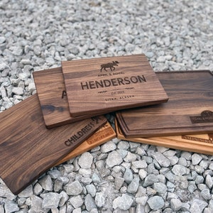 K&ouml;nnte beinhalten: F&uuml;nf verschiedene Schneidebretter aus Holz mit verschiedenen Gravuren. Die Bretter sind aus Walnussholz gefertigt und haben eine nat&uuml;rliche Holzoptik. Die Gravuren beinhalten Namen, Daten und Orte. Ein Brett hat ein Elch-Design.