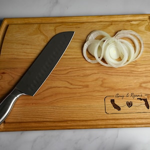 May include: A wooden cutting board with a rounded edge and a handle. The board is engraved with the text "Amy & Ryan's" and outlines of California and Florida with a heart in the middle. A silver knife and sliced onions are on the board.