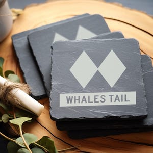 May include: Set of four black slate coasters with a white mountain peak design and the text "WHALES TAIL".