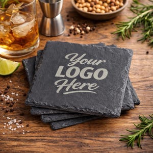 May include: A stack of four square, dark gray slate coasters with the text "Your LOGO Here" etched in a light gray font. The coasters are on a wooden surface, with a cocktail, lime, and garnishes in the background.