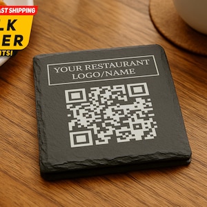 May include: A black slate coaster with a QR code and the text "YOUR RESTAURANT LOGO/NAME" in white. The coaster sits on a wooden table, near a plate with a croissant and a cup of coffee.