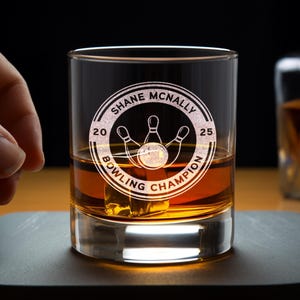 May include: A clear glass rocks glass with a personalized inscription that reads "Shane McNally 20 Bowling Champion 25". The inscription is in a circle with a bowling pin and ball graphic.