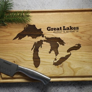 K&ouml;nnte beinhalten: Ein Schneidebrett aus Holz mit einem lasergravierten Design der Gro&szlig;en Seen. Das Brett ist hellbraun mit einem dunkelbraunen Rand. Der Text "Great Lakes 45.0522&deg; N, 82.4846&deg; W" ist in das Brett graviert.