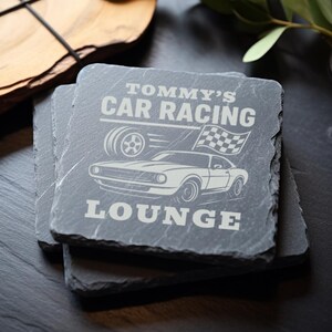 May include: A set of four black slate coasters with a white design. The top coaster features the text "Tommy's Car Racing Lounge" with a drawing of a car and a checkered flag.