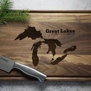 K&ouml;nnte beinhalten: Ein Schneidebrett aus Holz mit einer eingravierten Karte der Gro&szlig;en Seen. Das Brett ist braun und hat eine abgerundete Kante. Der Text "Great Lakes 45.0522&deg; N, 82.4846&deg; W" ist in das Brett graviert.