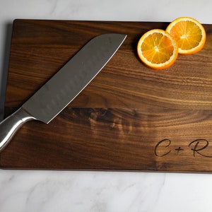 May include: A dark brown wooden cutting board with a silver knife and two orange slices. The board is engraved with the initials "C+R".