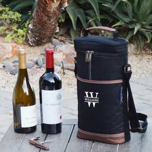May include: Black insulated wine tote bag with a brown leather handle and trim. The bag has a monogrammed initial "W" with the name "Williams" printed below. The bag is sitting on a wooden table with two bottles of wine and a corkscrew.