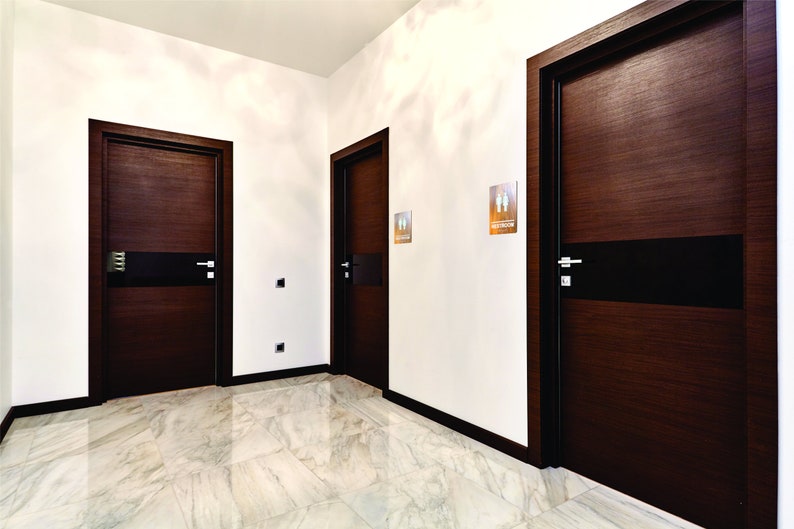 May include: Three dark brown wooden doors with black accents and silver door handles. The doors are in a hallway with white walls and a white marble floor.