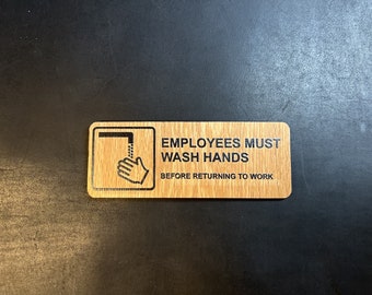May include: A wooden sign with the text "Employees Must Wash Hands Before Returning To Work". The sign has a black and white icon of a hand washing under a faucet.