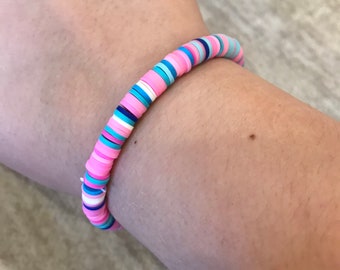 May include: A colorful bracelet made of pink, blue, and white clay beads. The bracelet is on a person's wrist.