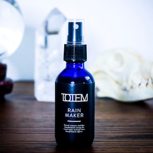 May include: A blue glass bottle with a black spray top and label that reads "TOTEM RAIN MAKER". The bottle is sitting on a wooden surface with a crystal and a white skull in the background.