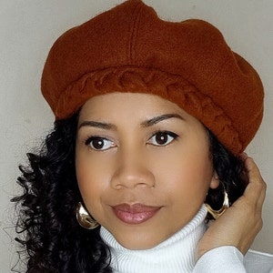Peut inclure: Une femme portant un béret marron avec un bord plié et de grandes boucles d'oreilles en or. Elle porte un pull à col roulé blanc.
