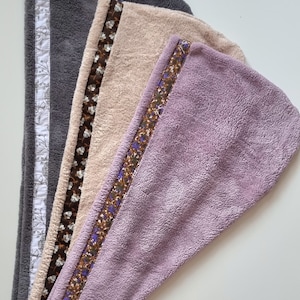 Pode incluir: Três toalhas de microfibra para cabelo em diferentes cores: cinza, bege e roxo. Cada toalha tem uma guarnição decorativa com um padrão floral.