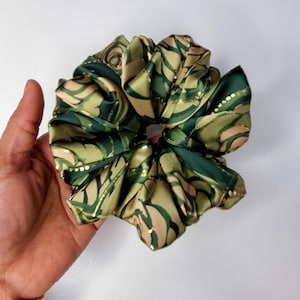 May include: A large, ruffled scrunchie in shades of green, tan, and gold. The fabric has a swirling, abstract pattern with gold accents. The scrunchie is held in a hand against a white background.