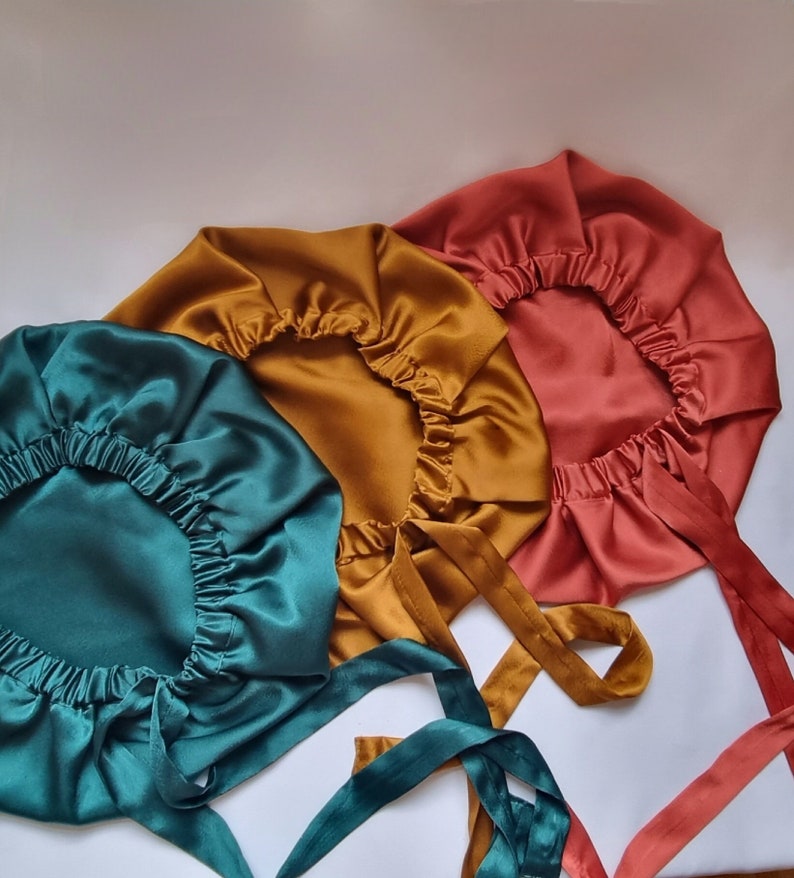 May include: Three satin bonnets in teal, gold, and coral. Each bonnet has a gathered elastic edge and long, matching ties. The bonnets are arranged on a white surface, showcasing their smooth, shiny texture and vibrant colors.