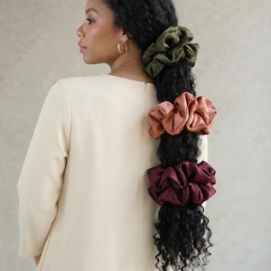 Op de afbeelding: Donker krullend haar van een vrouw is gestyled met drie grote satijnen scrunchies in olijfgroen, terracotta en bordeauxrood. De vrouw draagt een crèmekleurige jurk en gouden oorringen.