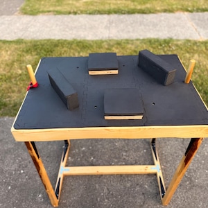 Pode incluir: Uma mesa profissional de queda de braço com estrutura de madeira, pernas anguladas e topo acolchoado preto. A superfície inclui duas almofadas quadradas para cotovelos, duas almofadas laterais retangulares e dois pinos verticais de madeira.
