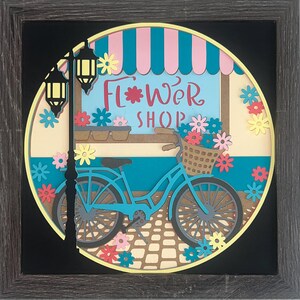 May include: A layered paper shadow box art piece in a dark wood frame featuring a circular scene of a blue bicycle with a flower-filled basket in front of a shop with a pink and blue striped awning and the text Flower Shop in red script.