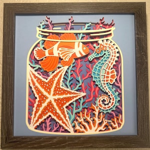 May include: A multi-layered paper art piece in a dark wood frame depicting an underwater scene inside a glass jar. The design features an orange clownfish, a turquoise and orange seahorse, and a large orange starfish surrounded by coral.