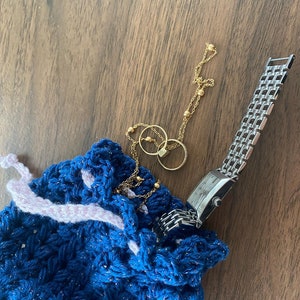 Könnte beinhalten: Eine blaue gehäkelte Kordeltasche mit rosa Besatz, die eine goldene Kettenkette mit zwei Ringen und eine silberne Uhr enthält.