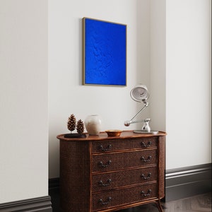 May include: A dark brown wooden dresser with four drawers and a woven rattan front. The dresser has a curved front and sits on a dark wood floor. A small blue painting hangs on the wall above the dresser. A lamp with a silver shade sits on the dresser.