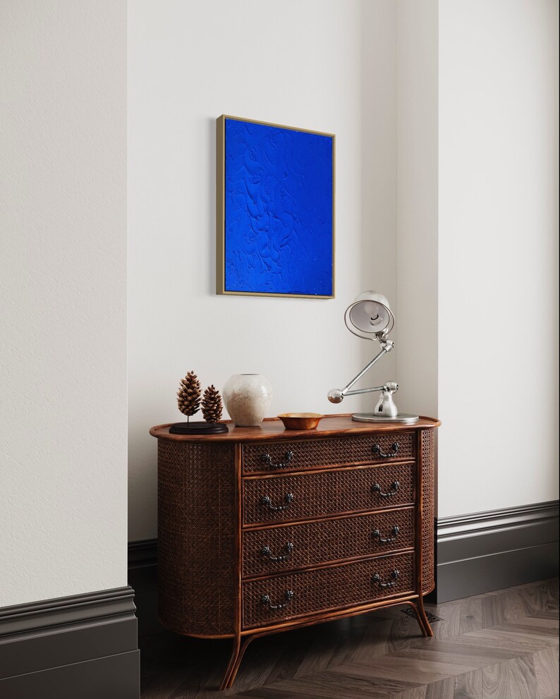 May include: A dark brown wooden dresser with four drawers and a woven rattan front. The dresser has a curved front and sits on a dark wood floor. A small blue painting hangs on the wall above the dresser. A lamp with a silver shade sits on the dresser.