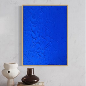 May include: A framed abstract painting with a textured blue background. The painting is in a gold frame and is hanging on a white wall.