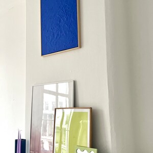May include: A white wall with a blue framed painting, a white framed mirror, a green framed poster, a small mirror with a wavy frame, a silver disco ball, and a glass vase on a black marble mantelpiece. The poster says "Pantoni A-SOCIAL MEDIA".