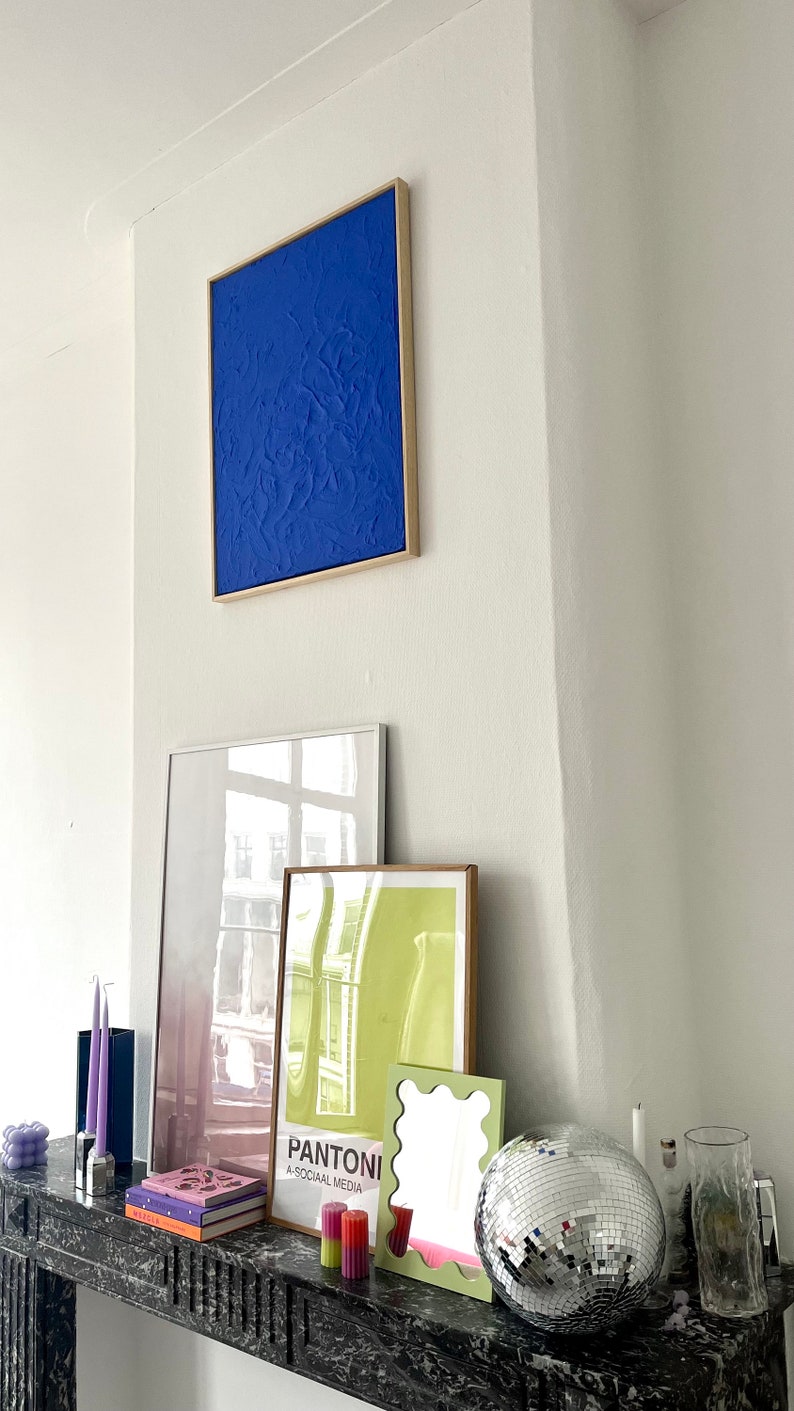 May include: A white wall with a blue framed painting, a white framed mirror, a green framed poster, a small mirror with a wavy frame, a silver disco ball, and a glass vase on a black marble mantelpiece. The poster says "Pantoni A-SOCIAL MEDIA".