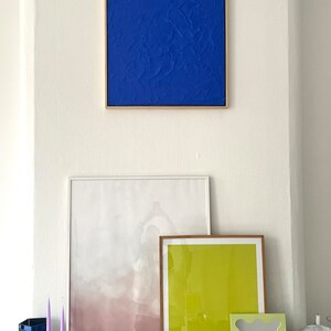 May include: A white wall with three framed pictures and a disco ball. The pictures are a blue abstract painting, a white and pink abstract painting, and a yellow and green abstract painting. The bottom of the wall has a black marble surface with two purple candles, a blue glass vase, and a book titled "Mezcla".