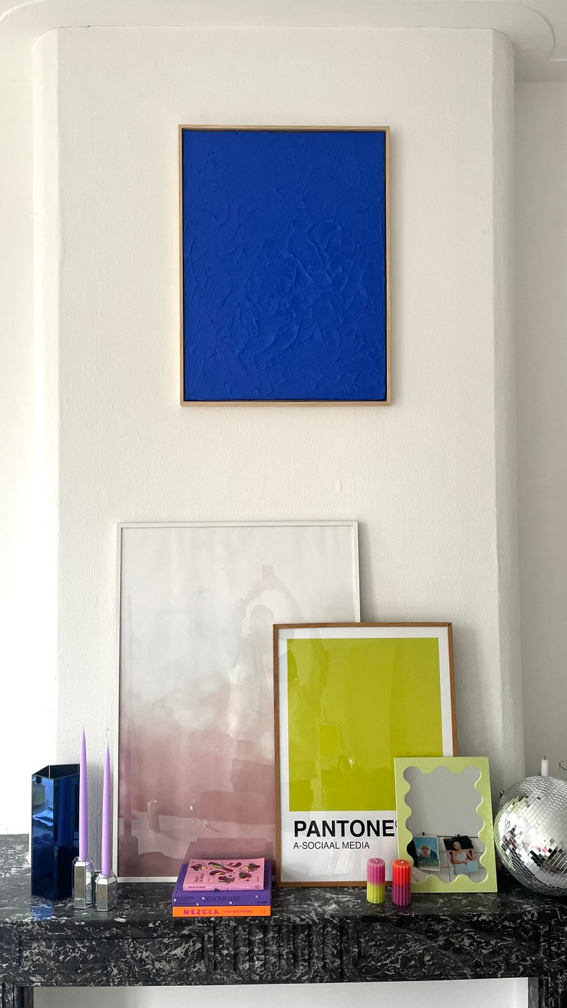 May include: A white wall with three framed pictures and a disco ball. The pictures are a blue abstract painting, a white and pink abstract painting, and a yellow and green abstract painting. The bottom of the wall has a black marble surface with two purple candles, a blue glass vase, and a book titled "Mezcla".
